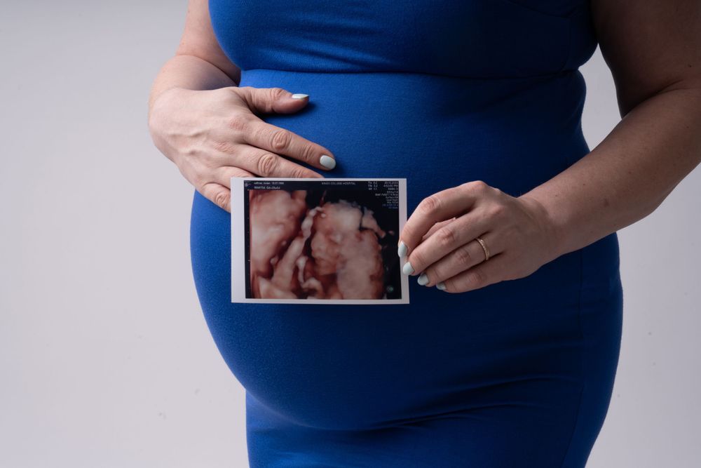 Expectant couple in blue dress and casual wear holding ultrasound photos during pregnancy shoot.