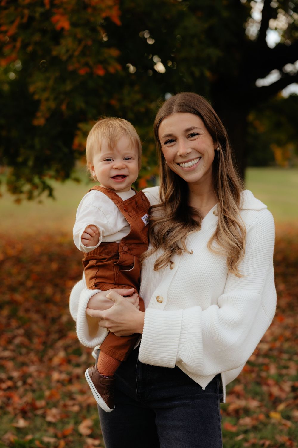 Parent and baby in matching white and brown outfits playing together among fall leaves.