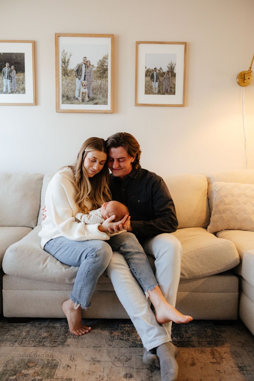 New parents lounge together on a gray couch in a cozy living room with their newborn.
