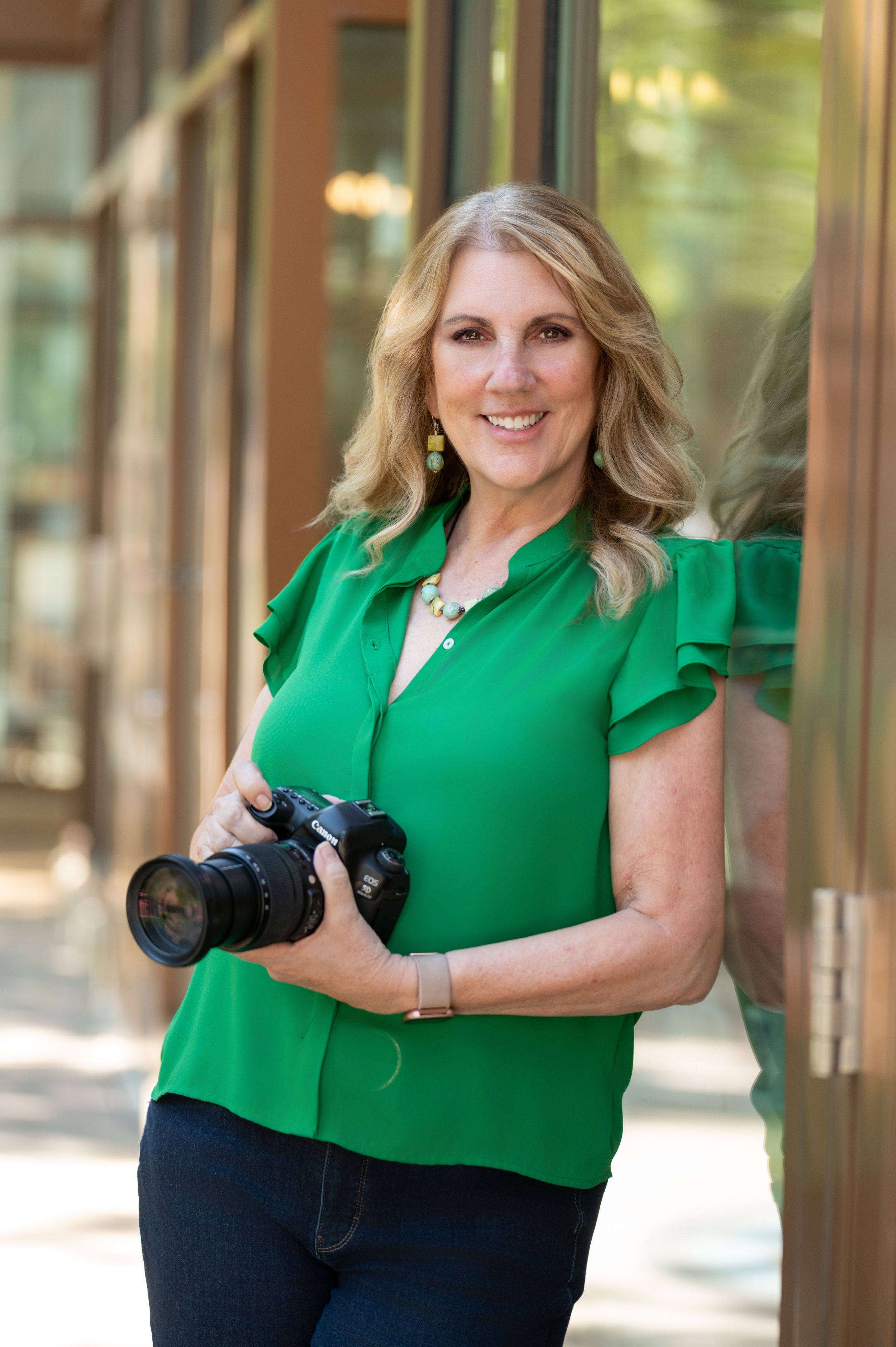 Professional photographer wearing bright green blouse holds DSLR camera while posing against reflective window background.