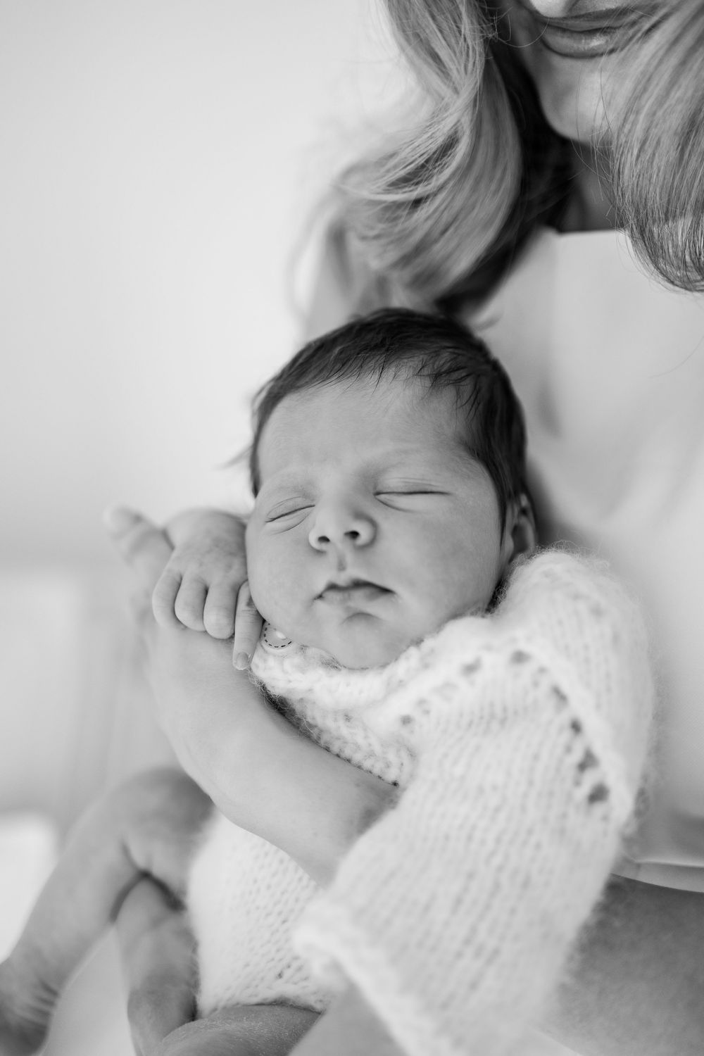 A peaceful sleeping newborn baby wrapped in a white knit blanket being held in loving arms.