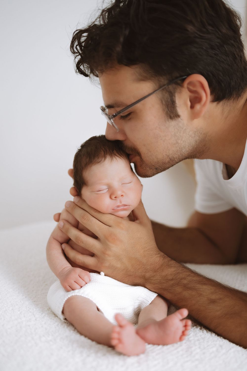A tender moment between parent and sleeping newborn baby on white blanket in soft natural light.