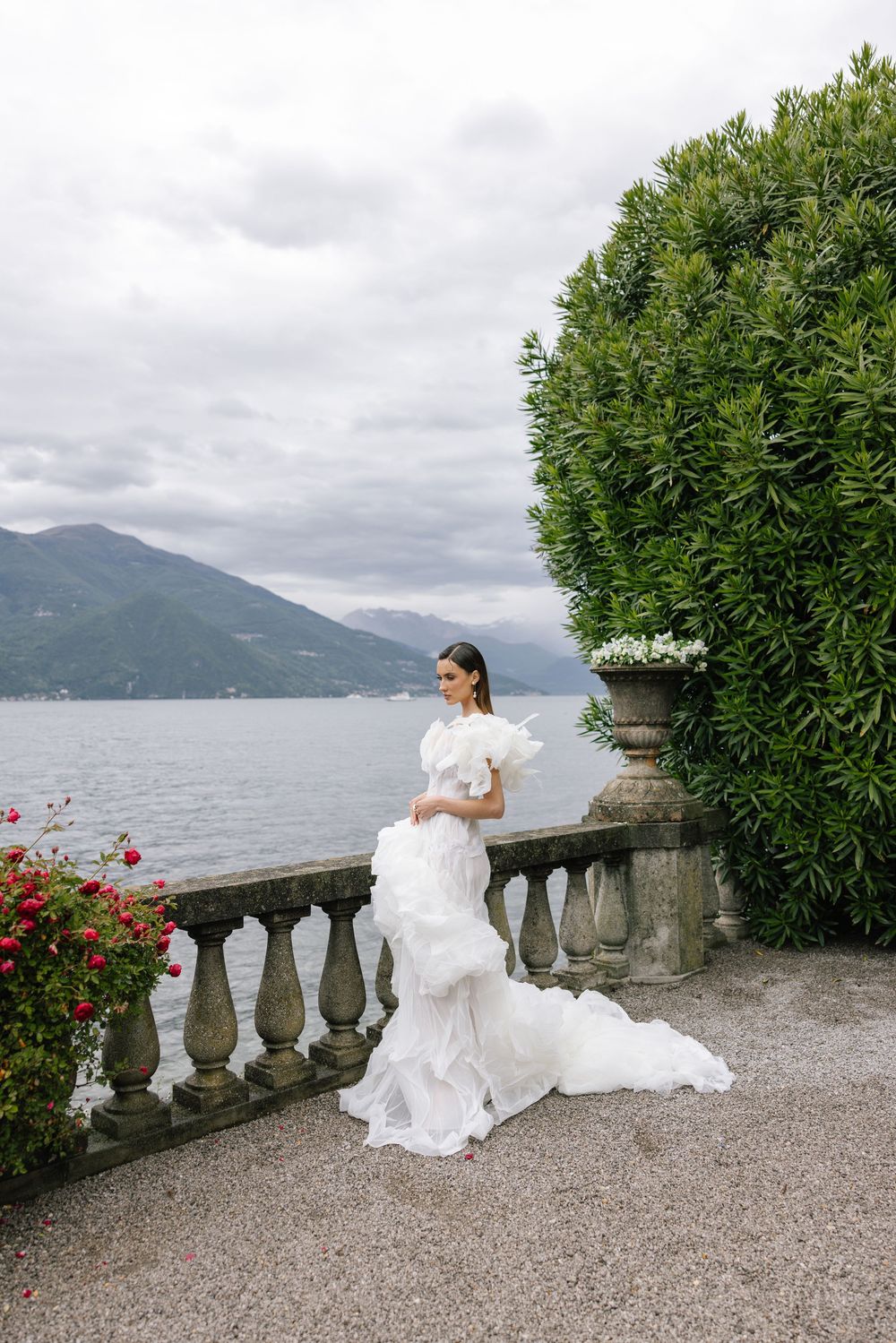 A bride in a flowing white dress sits on a stone balustrade overlooking Lake Como and mountains in Italy.