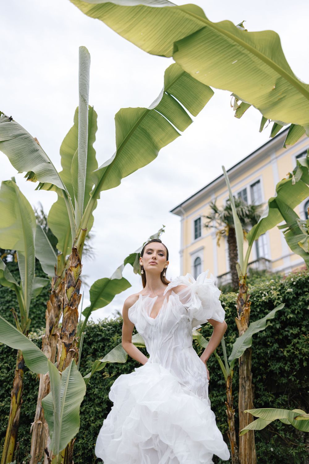 A model in a flowing white dress poses elegantly among tropical banana leaf plants in a garden setting.