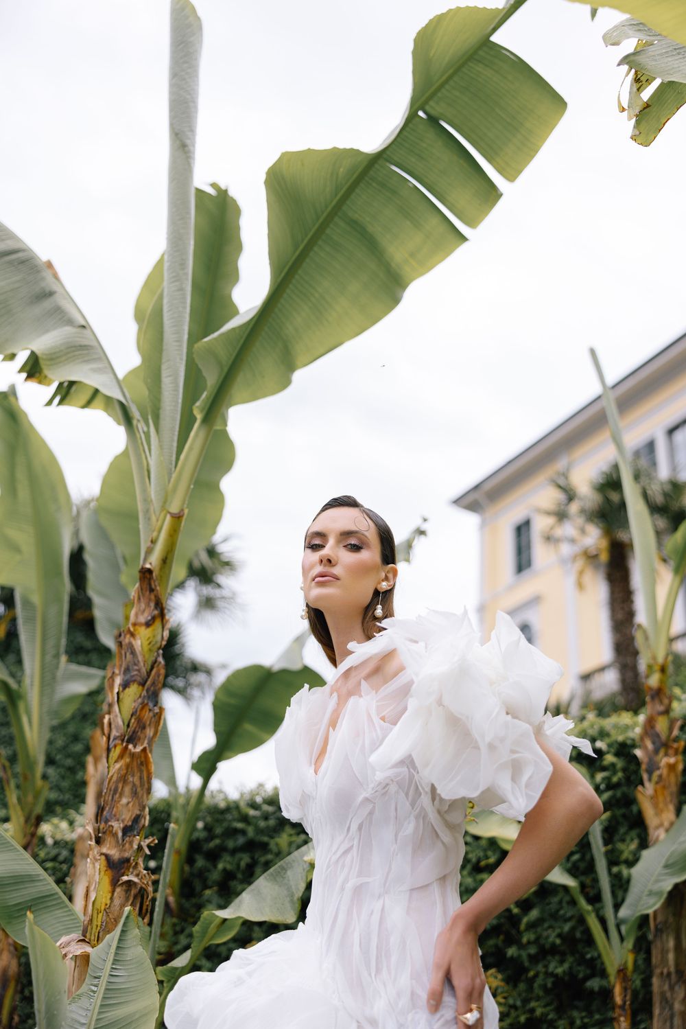Model in flowing white dress poses gracefully under tropical banana leaves in an outdoor garden setting.