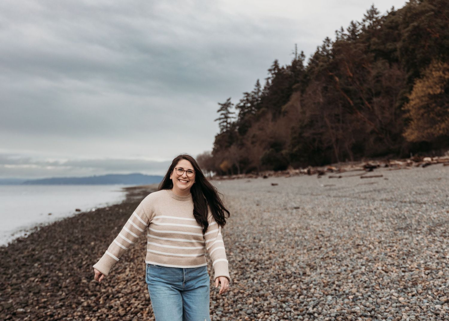 A person walks along a rocky beach shoreline with evergreen trees on the cliffside under cloudy skies.
