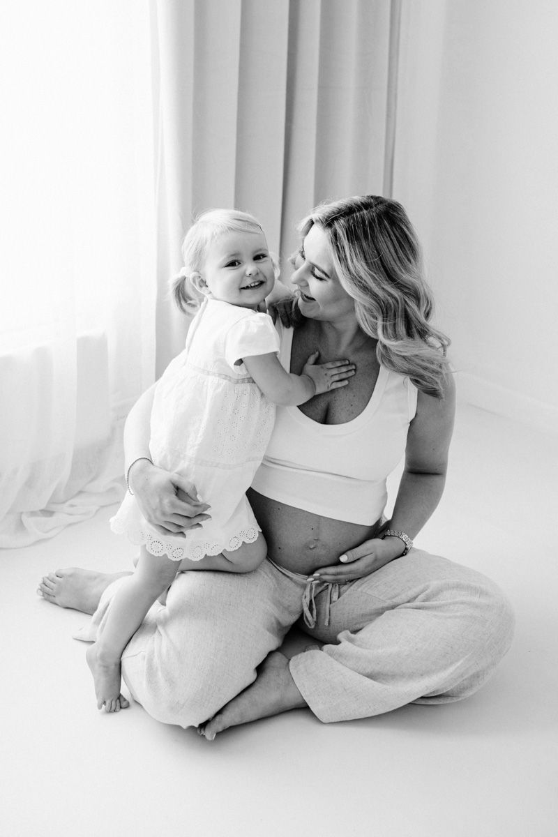 Black and white portrait of a pregnant woman in casual attire sitting with a toddler in a bright, minimalist setting.
