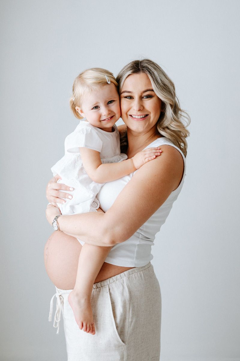 A tender portrait of a parent and baby in white clothing embracing against a light gray background.