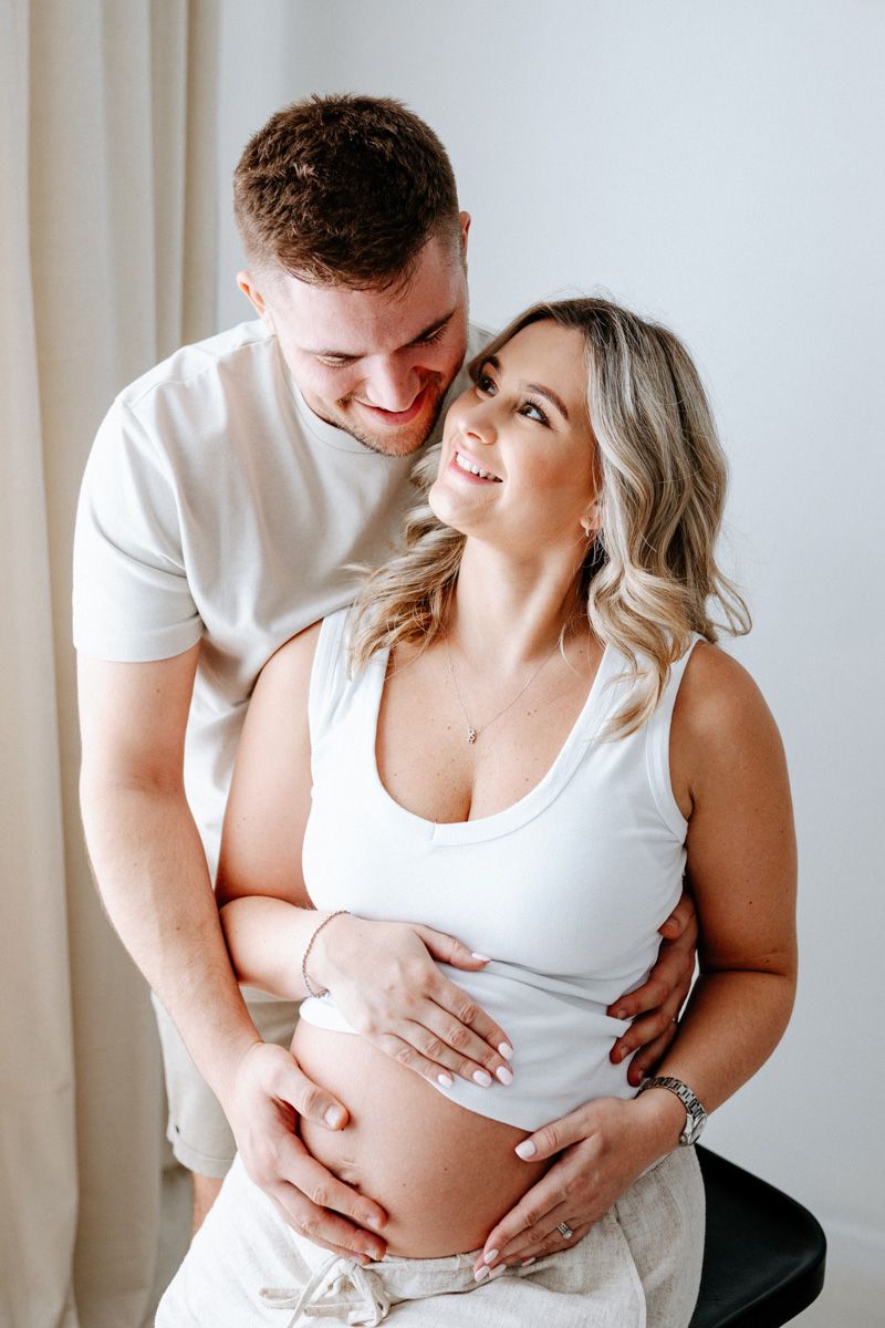 A couple in white shirts share an intimate maternity photoshoot moment against a light background.
