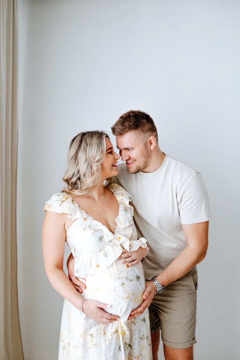 Expecting couple shares intimate moment in white floral dress and casual attire against neutral wall.