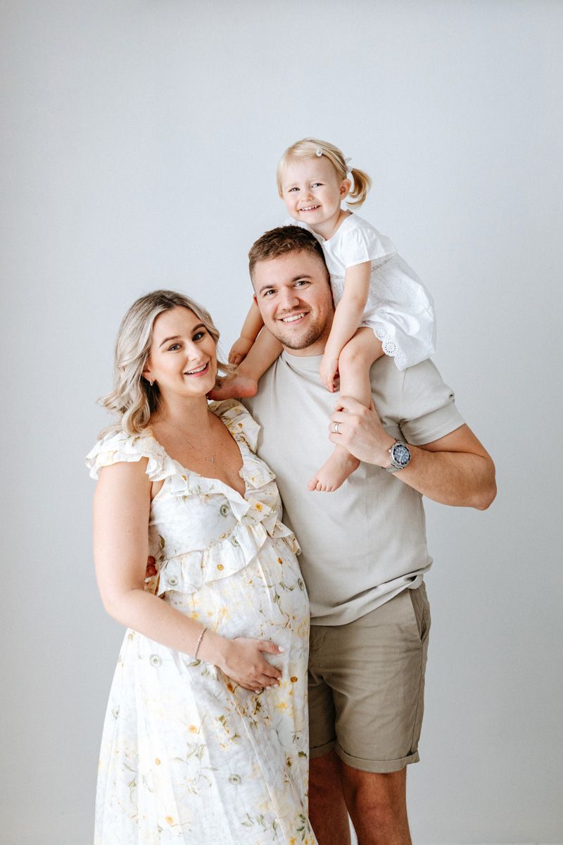 A family in white and beige outfits poses together against a light gray studio backdrop.