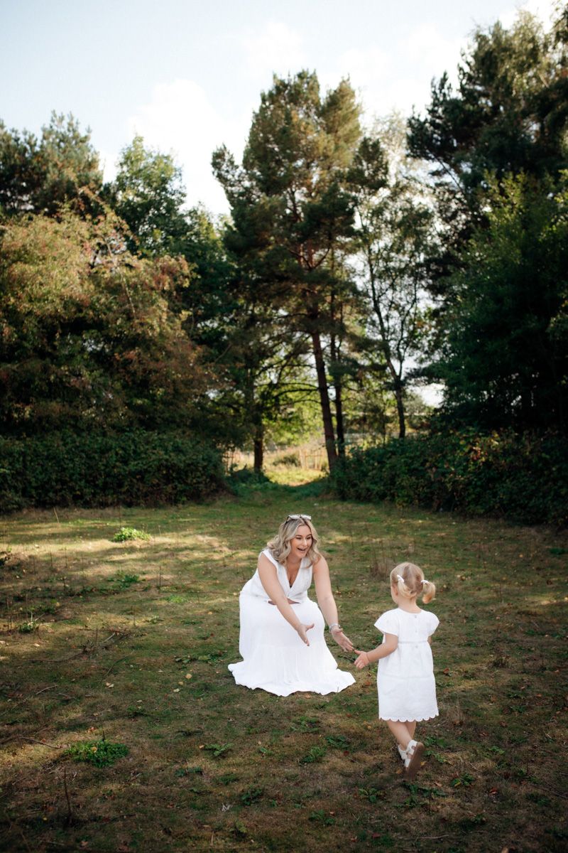 Two figures in white dresses share a tender moment in a sunlit forest clearing surrounded by tall pine trees.