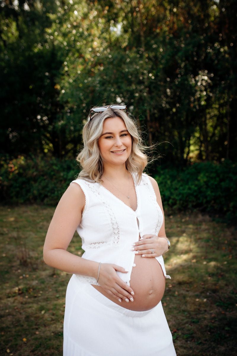 A maternity photoshoot in a white dress against a natural outdoor backdrop with trees and foliage.