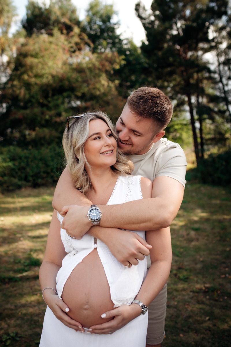 A couple embraces in a tender maternity photoshoot outdoors in a natural setting with trees in the background.
