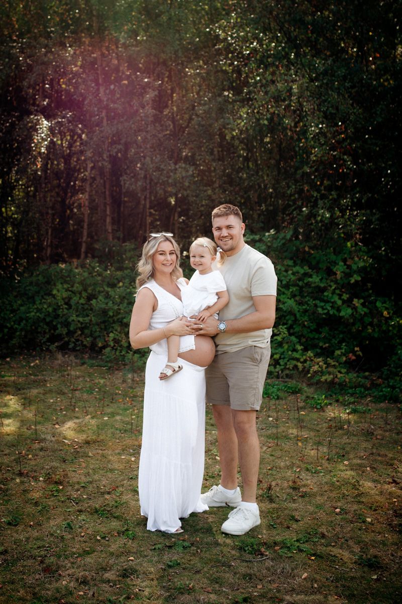 A family wearing white outfits poses together in a wooded outdoor setting during golden hour.