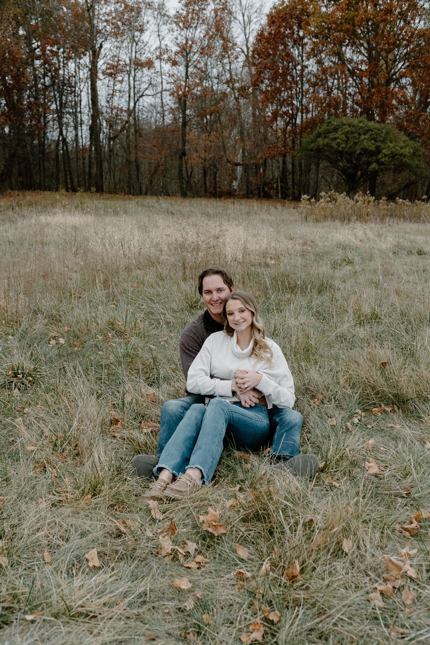 A couple sits together in a fall meadow surrounded by autumn trees with brown and orange foliage.