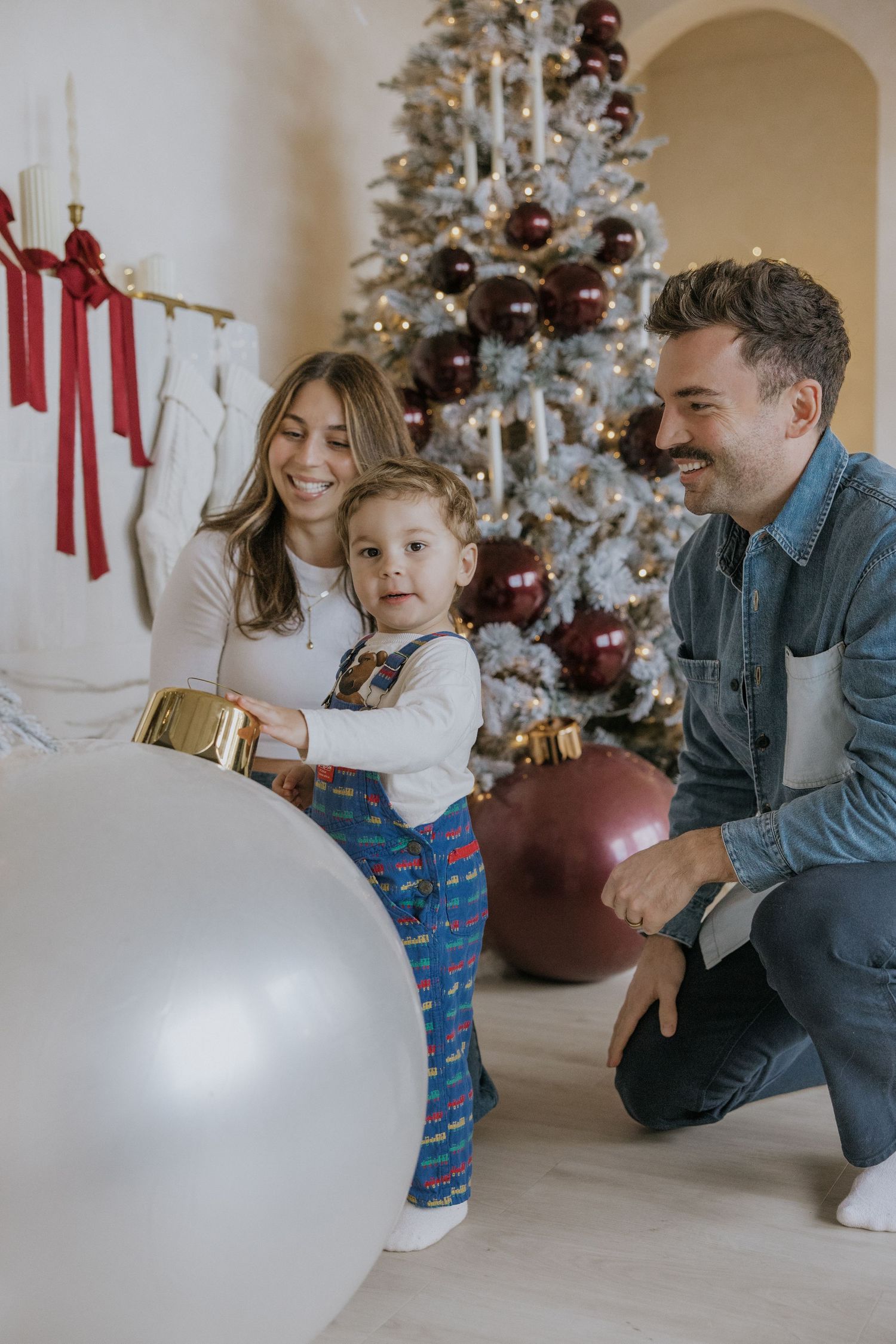 A family poses together by a decorated white Christmas tree with large ornament balls on the floor.