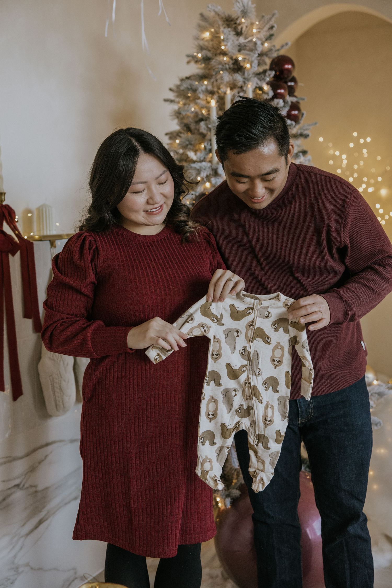 Couple in matching burgundy sweaters holding up a brown and white patterned baby onesie together.