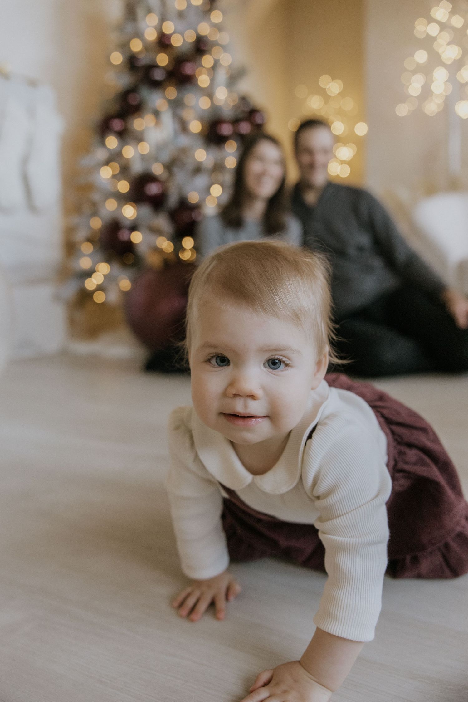 Baby in white sweater crawls on light floor with Christmas tree and twinkling lights in background.