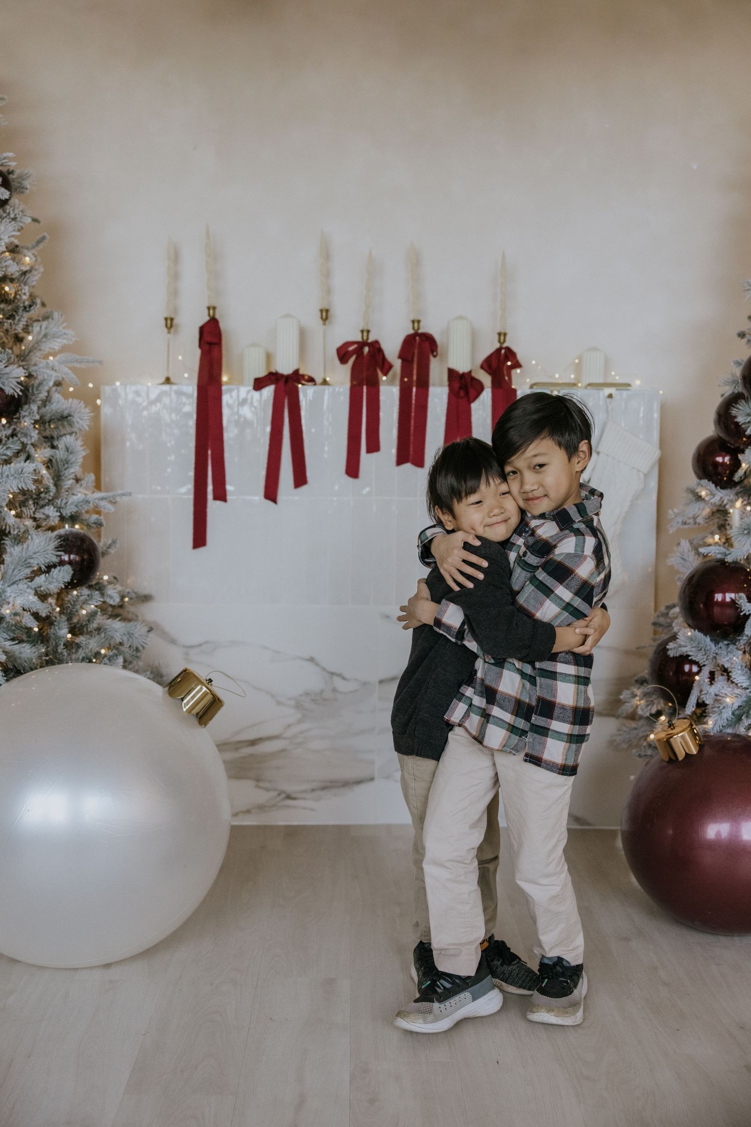 Two children in matching outfits play and laugh together near a white Christmas tree with red ornaments and decorations.
