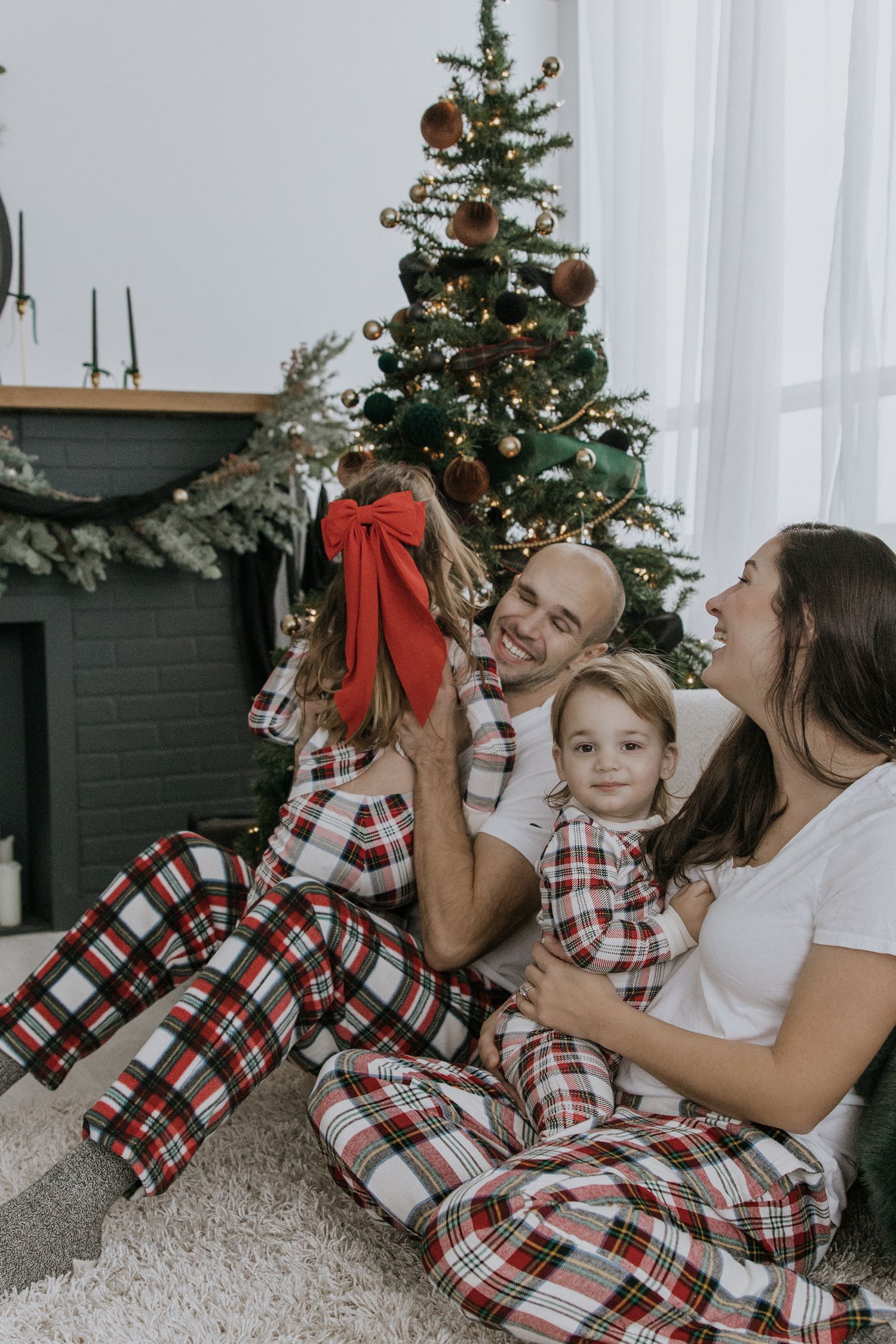Family wearing matching plaid pajamas cuddles together by decorated Christmas tree in living room.