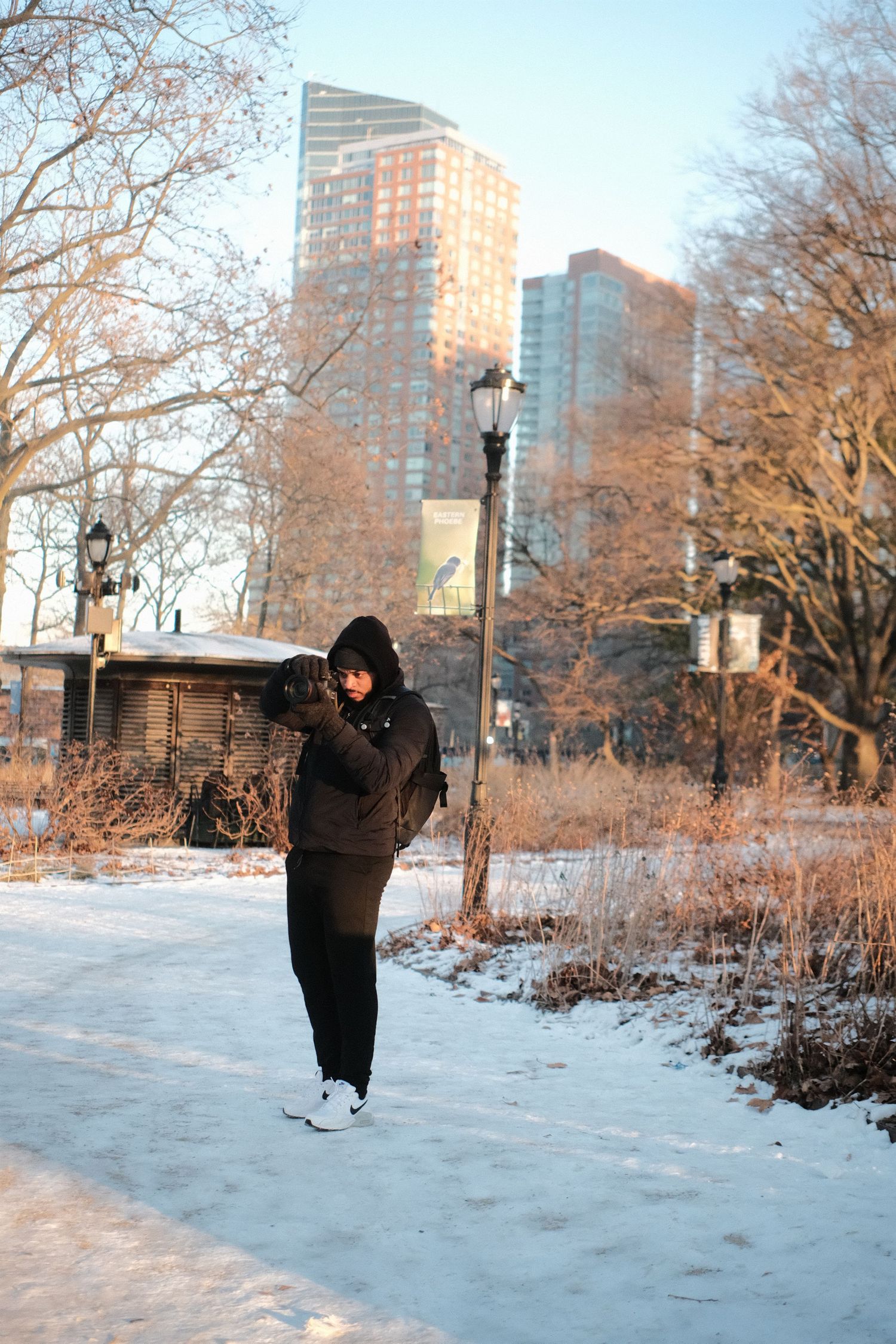 A person in winter clothing walks along a snowy path in an urban park with tall buildings in the background.