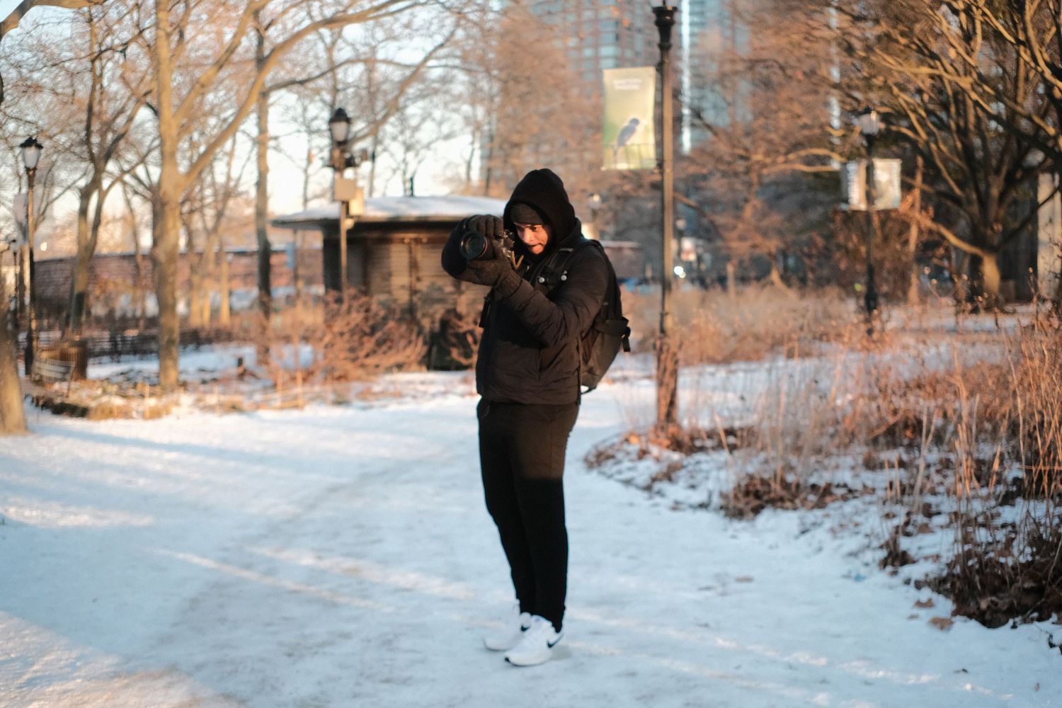 Figure in black winter attire strolls through a snow-covered urban park during golden hour.