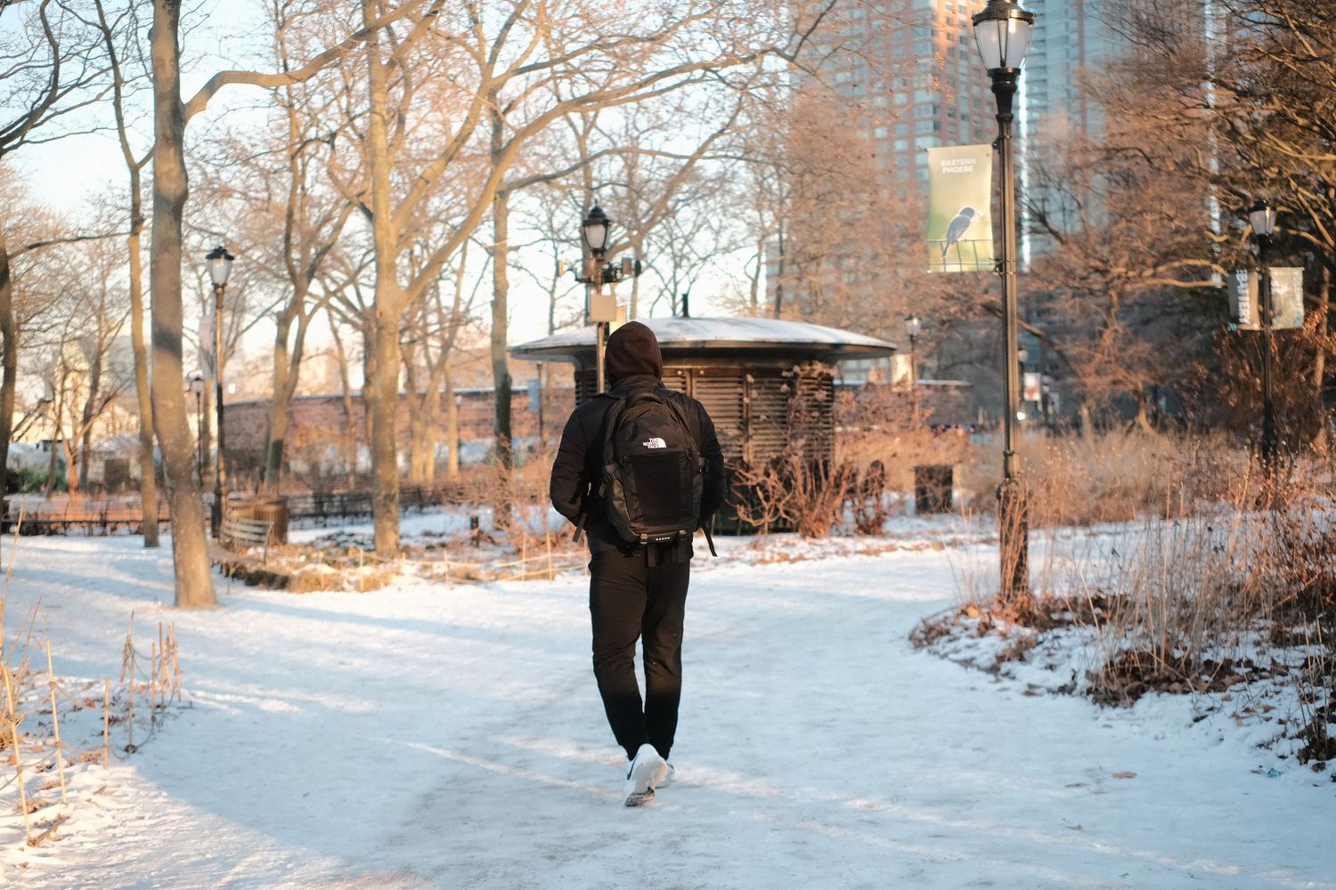 Person wearing dark clothing walks on snowy path with city buildings and bare trees in background.