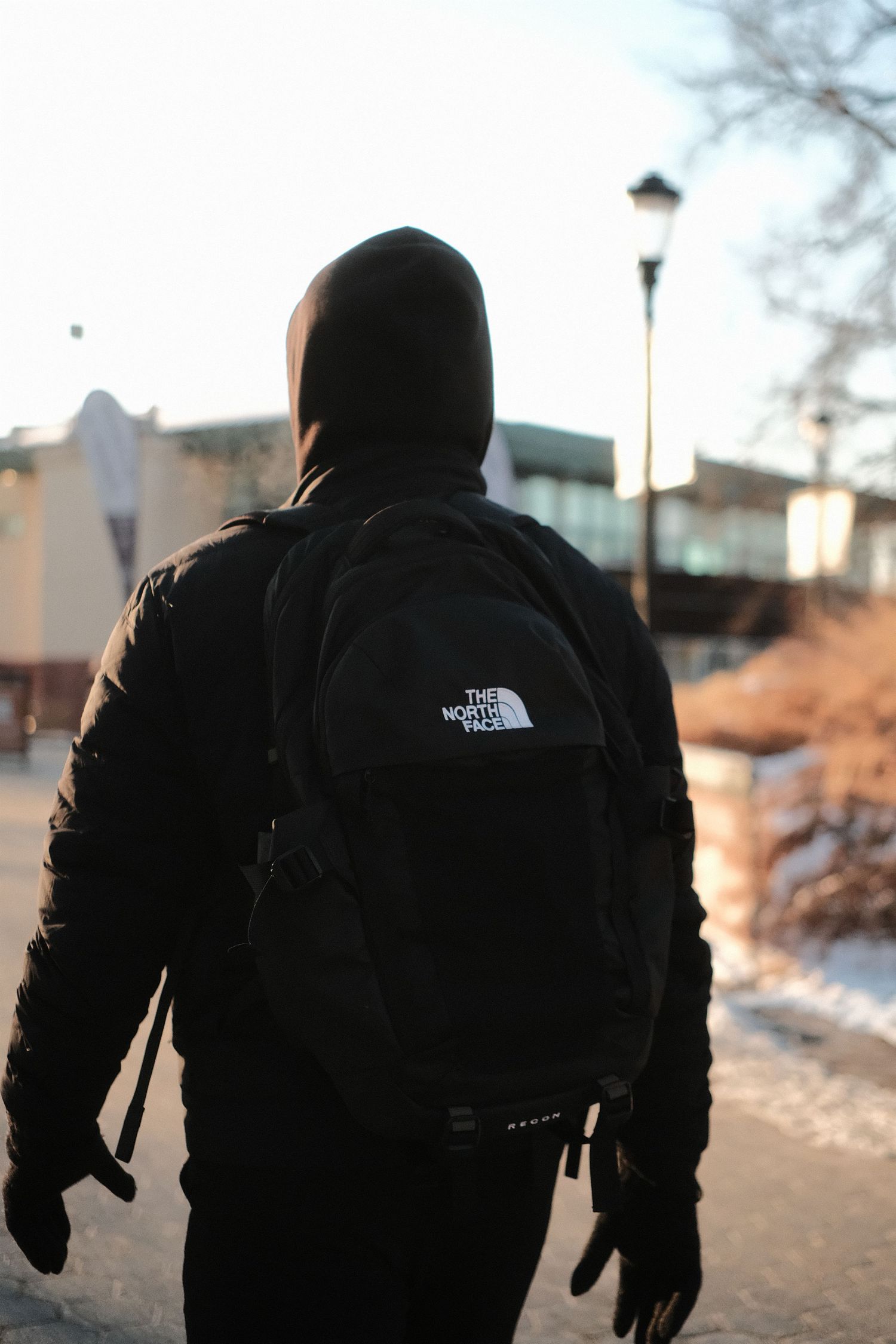 Person wearing black North Face jacket and backpack photographed from behind on urban path.
