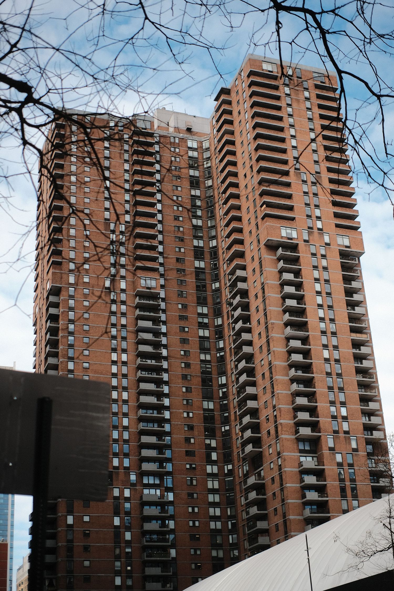Tall brick residential tower with balconies framed by bare tree branches against blue sky.