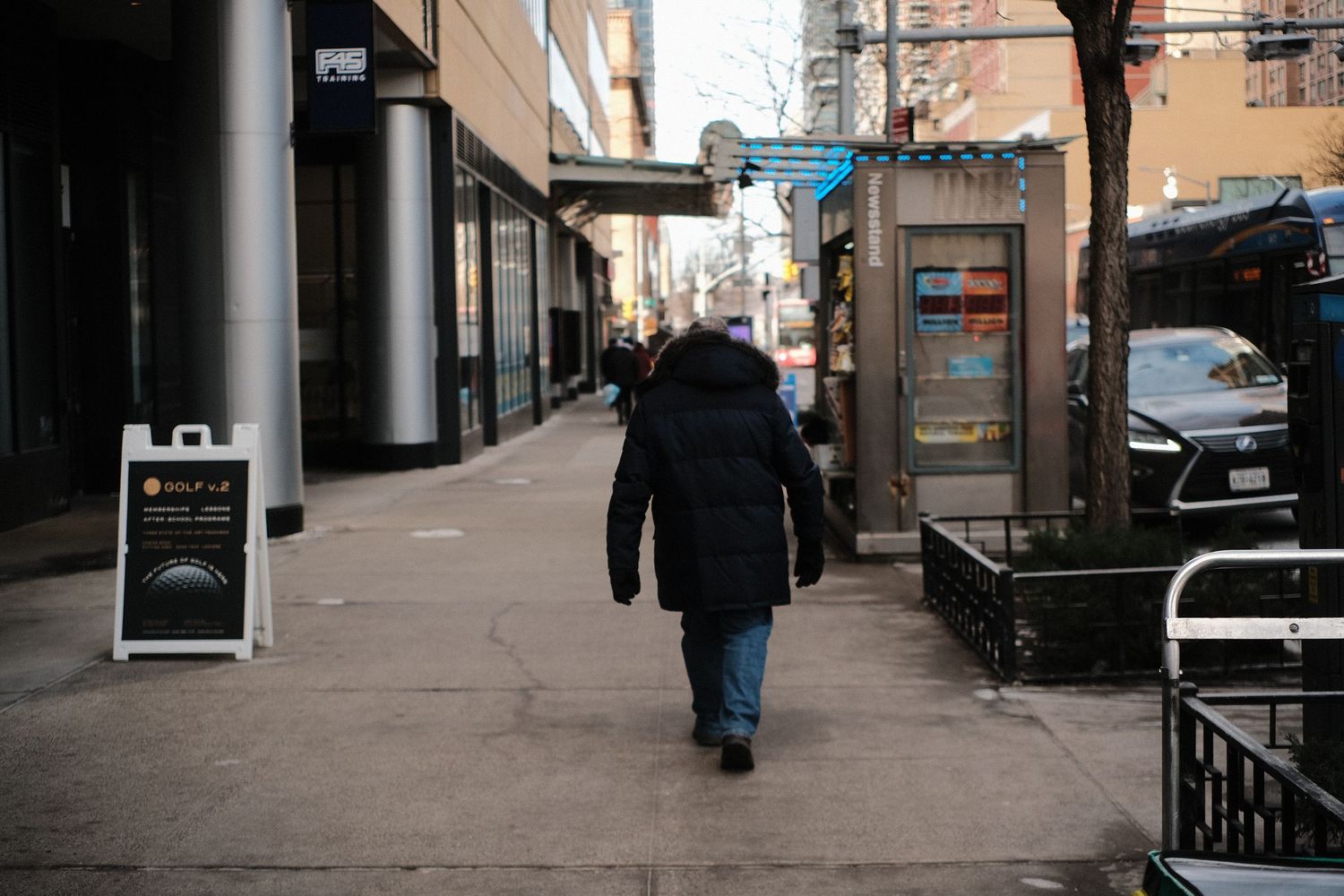 Urban street scene with pedestrians walking past brick buildings and storefronts on a city sidewalk.