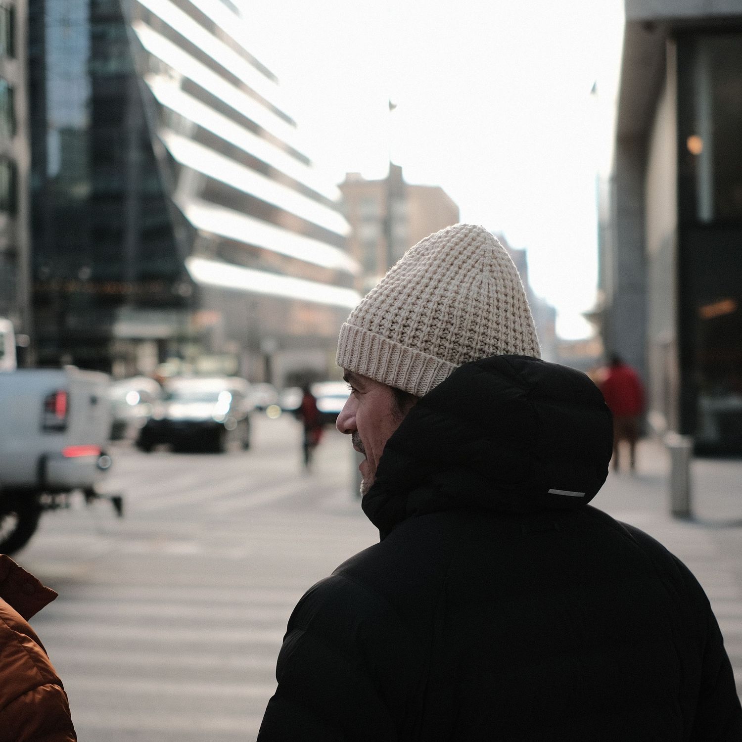 Person wearing a knit beanie and winter coat walking on an urban street with modern buildings.