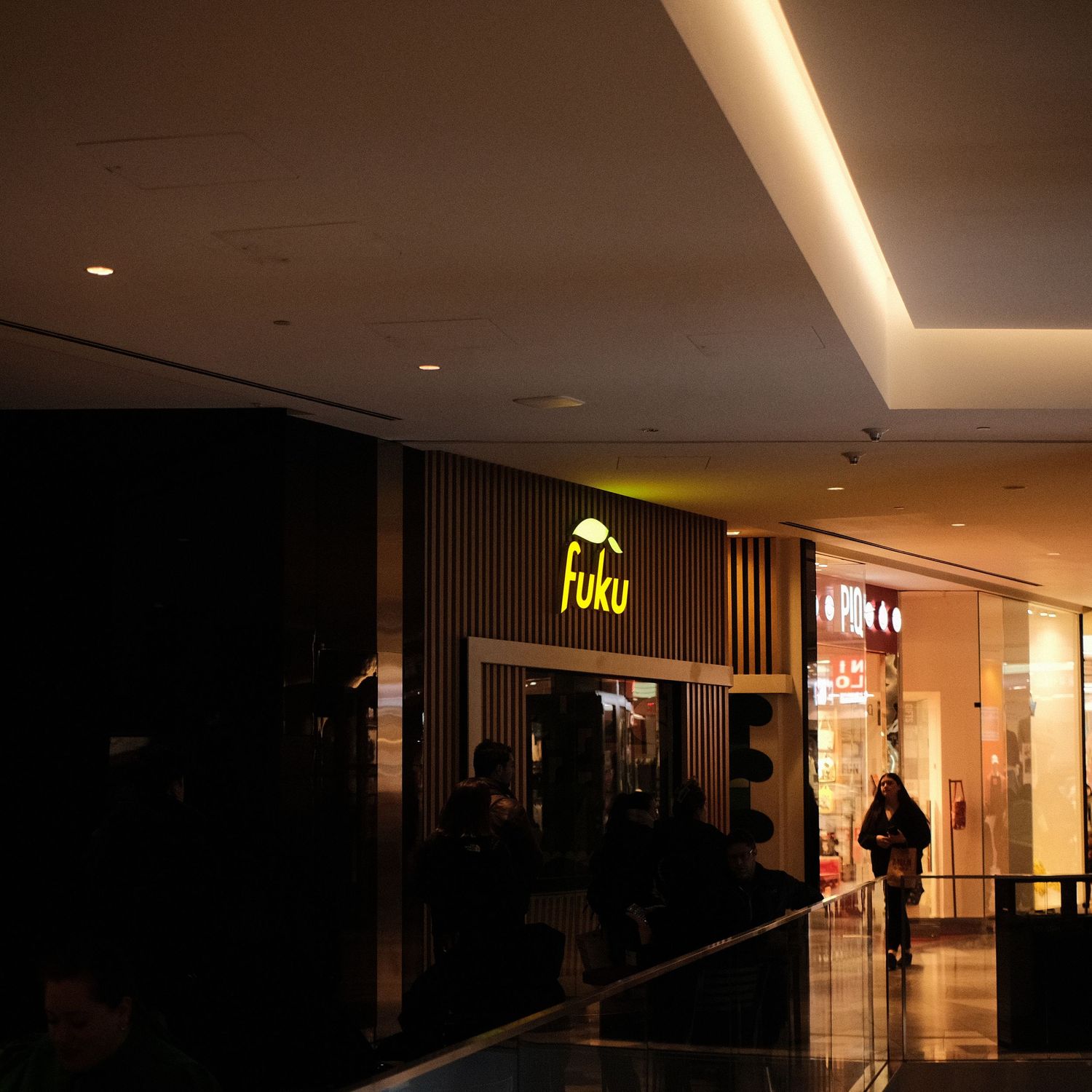Dimly lit shopping mall corridor with illuminated store signs and people walking.
