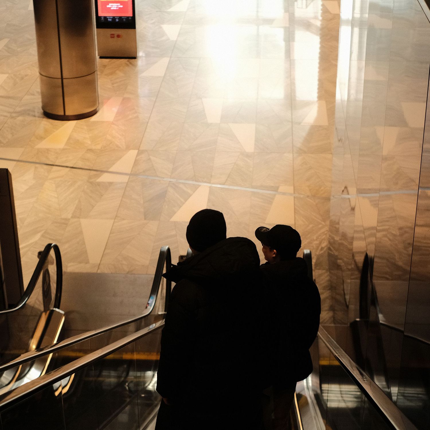 Silhouettes of people descending on an escalator in a brightly lit shopping mall.