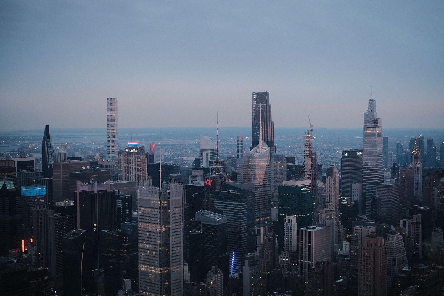 Panoramic cityscape view of Manhattan skyline during dusk with skyscrapers silhouetted against blue sky.