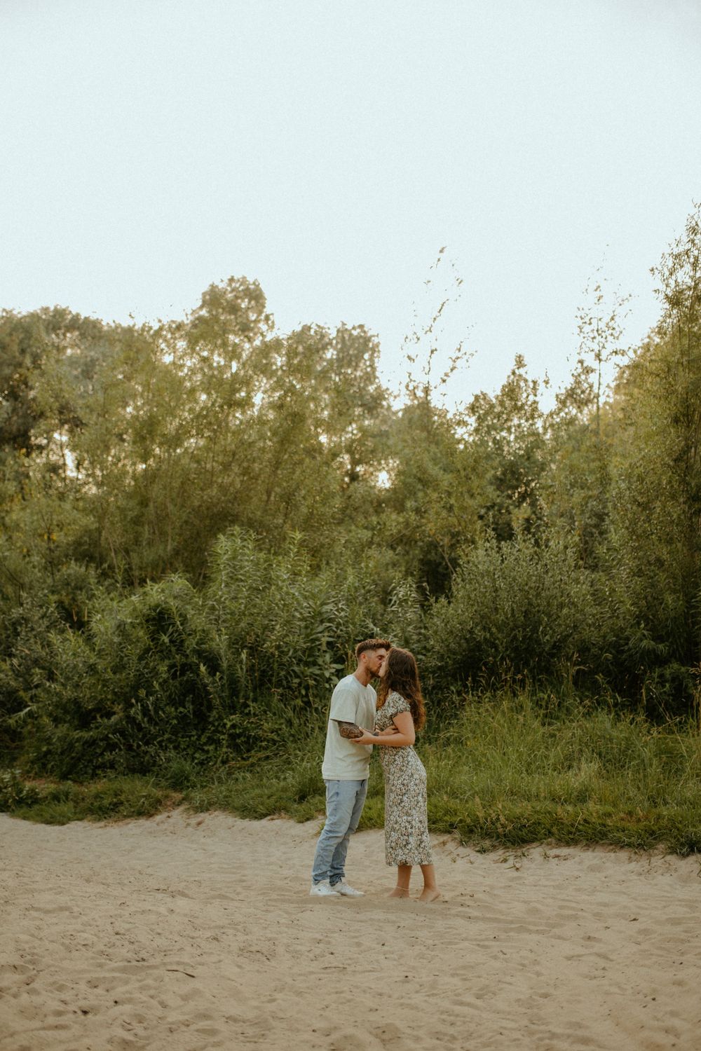A romantic couple embraces on a sandy beach path surrounded by green foliage during golden hour.