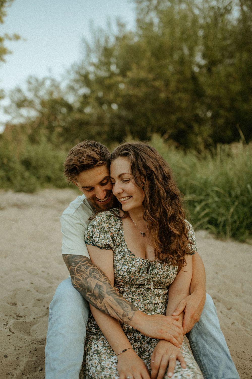 Intimate moment between a couple sitting on sand, wearing casual clothes against a natural backdrop.