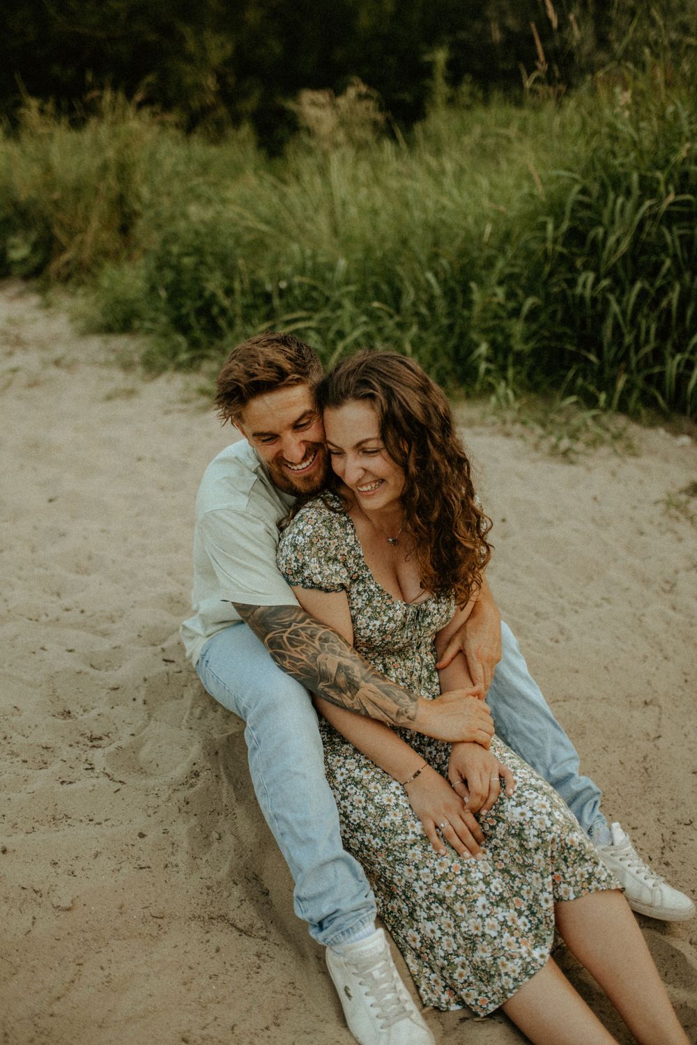 Couple in casual attire sharing intimate moments on sandy ground with lush vegetation behind them.
