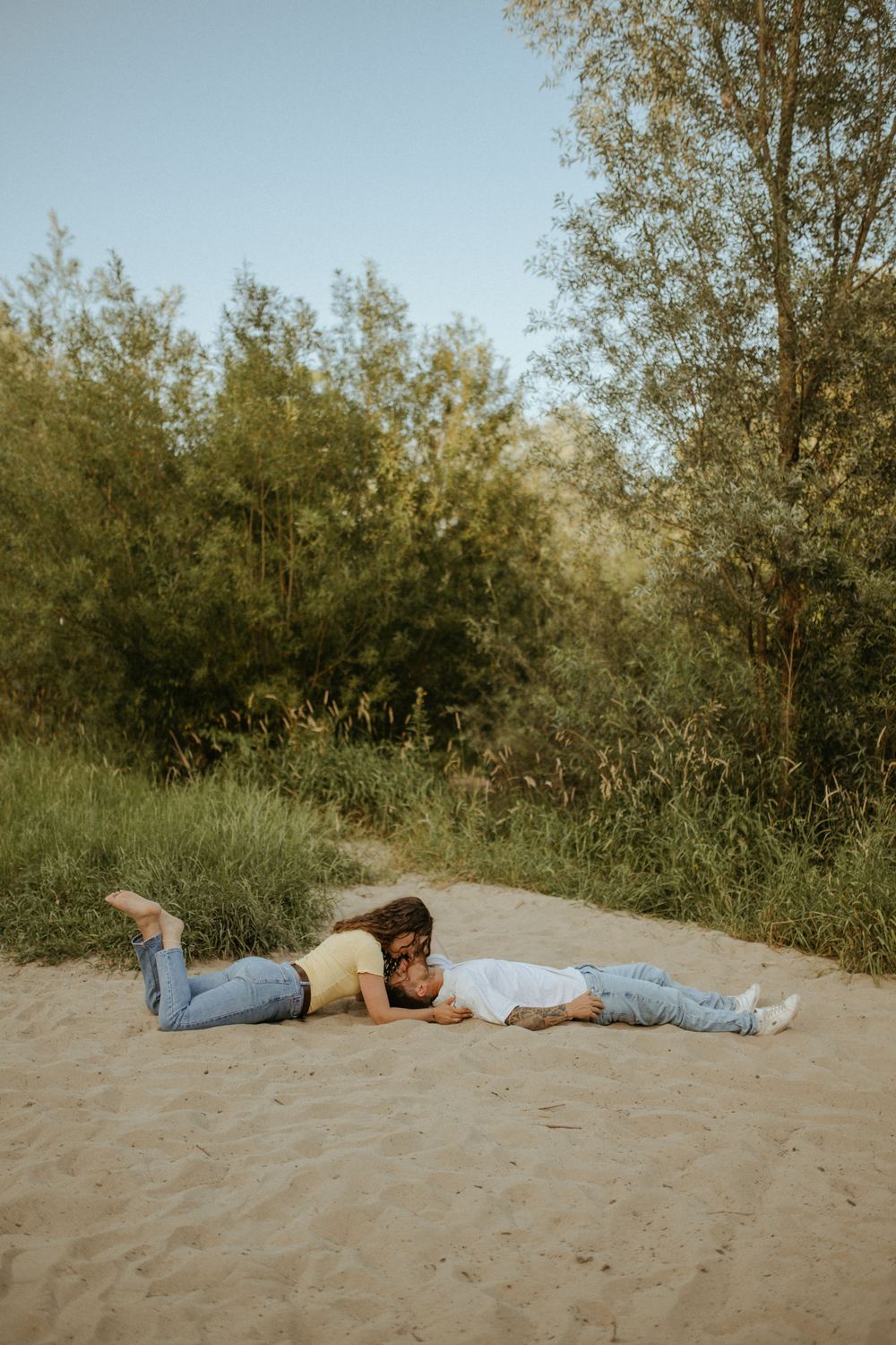 Two people lying down on sandy ground with trees and greenery in the background.