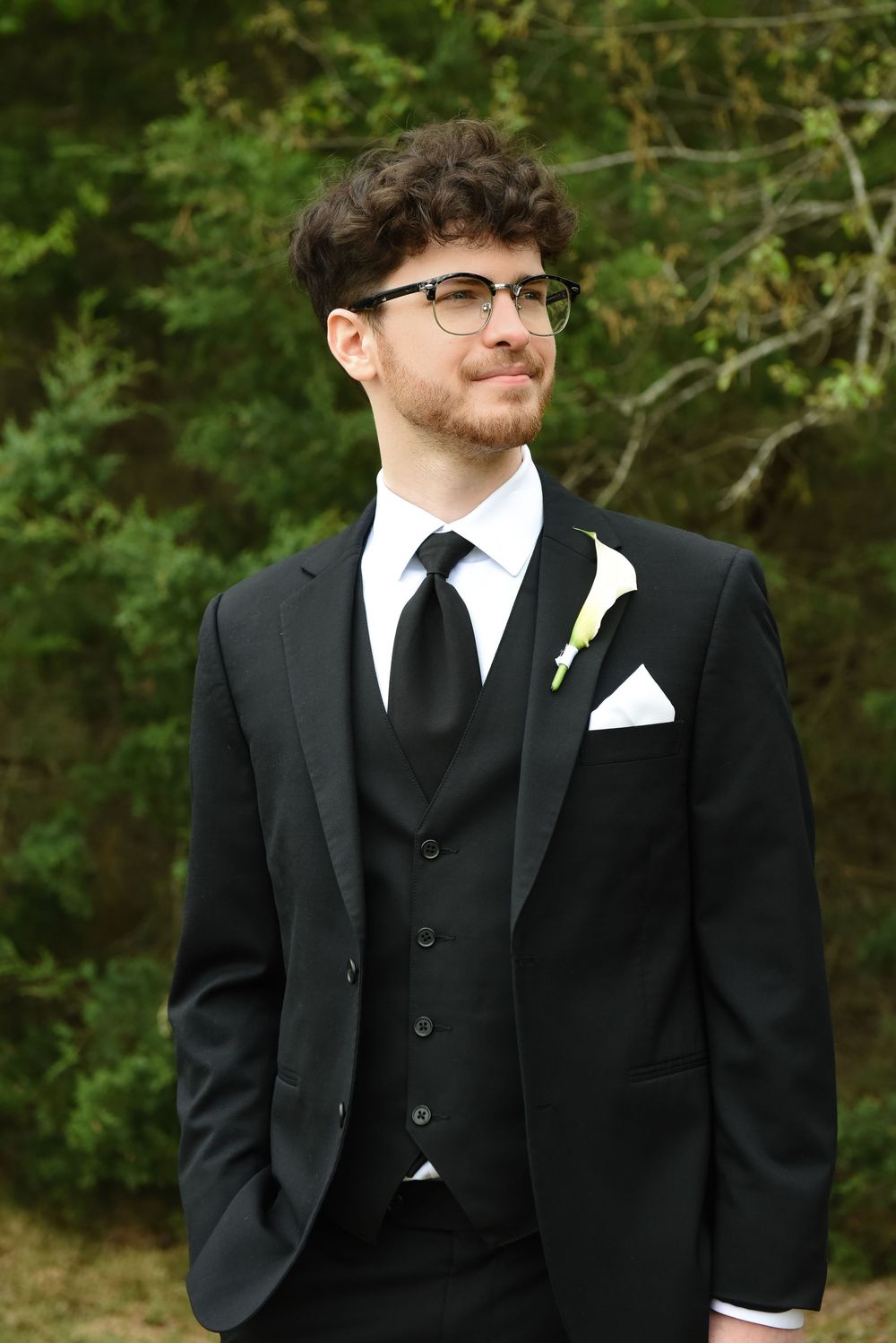Young man in black three-piece suit with tie and boutonniere, wearing glasses, posing outdoors at a formal event.