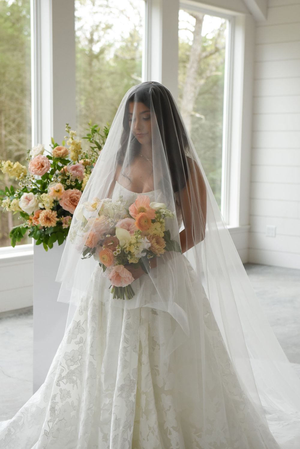 Bride in lace ball gown and sheer veil holds peach floral bouquet beside large floral arrangement in bright white room.