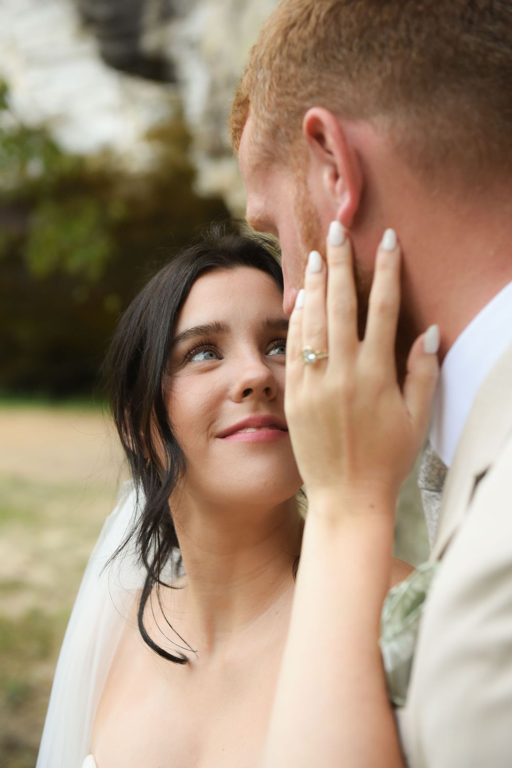 A bride reaches up to tenderly caress someone's face while wearing a wedding ring and white veil.
