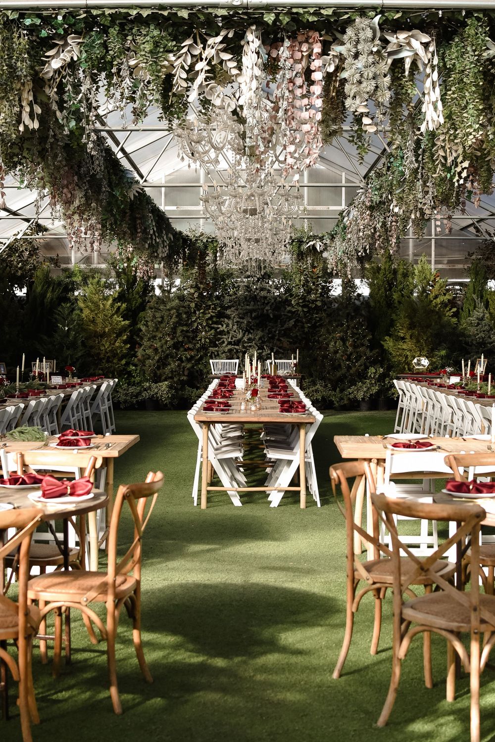 Elegant greenhouse wedding reception with floral chandeliers, long wooden tables, red napkins, and lush greenery backdrop.