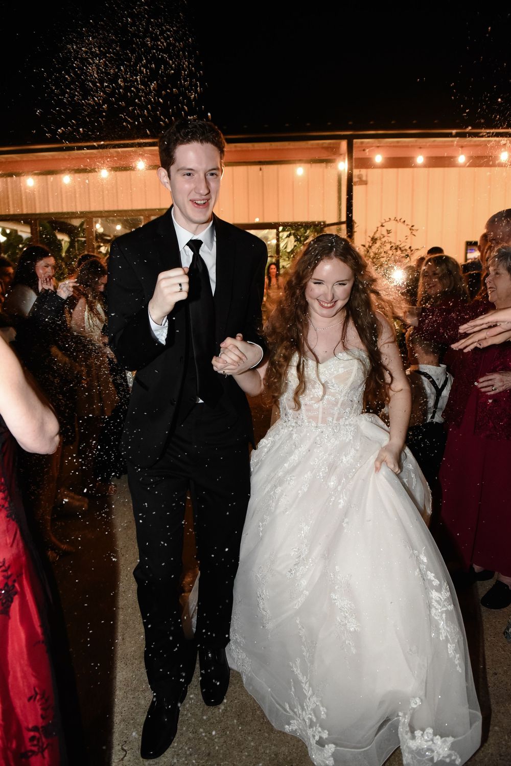 Joyful newlyweds exit their wedding reception, bride in lace gown and groom in black suit, surrounded by cheering guests.