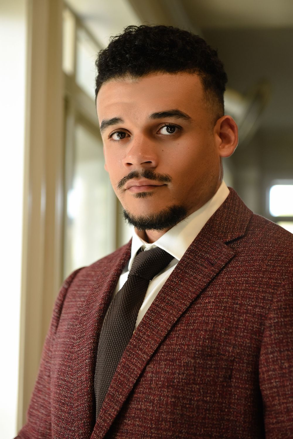 Young man in burgundy tweed blazer and dark tie poses confidently in professional portrait setting.