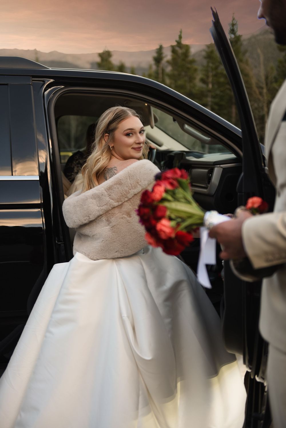 A person in a white winter wedding dress and fur shawl is helped into a car while holding red roses.