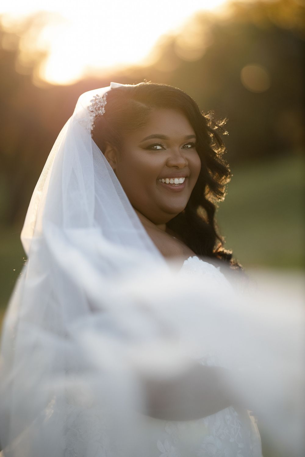 Radiant Black bride smiling in white veil and gown, golden sunset backlight glowing behind her outdoors.