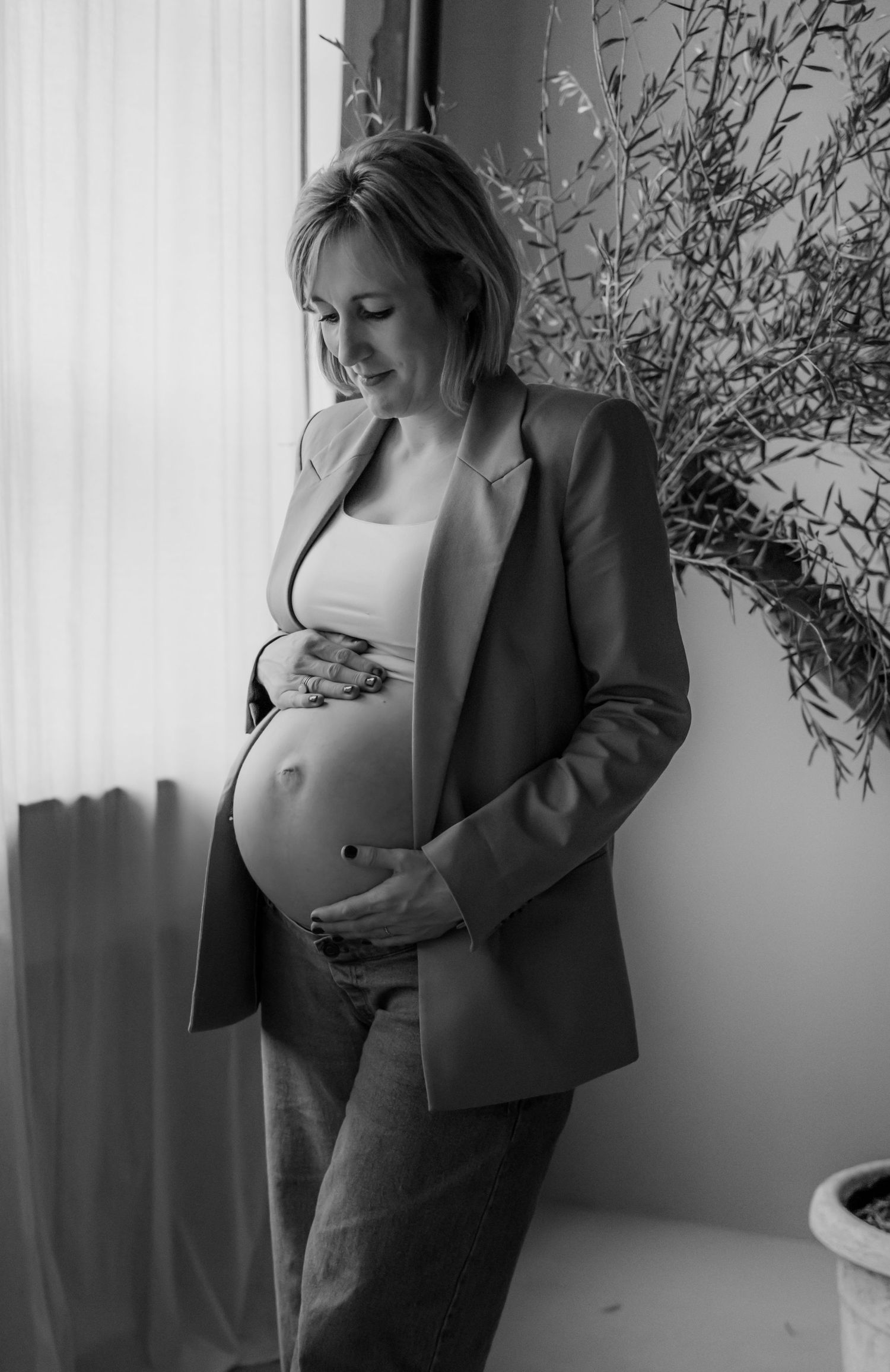 A maternity photoshoot featuring a pregnant woman in a blazer standing near a window with natural light.