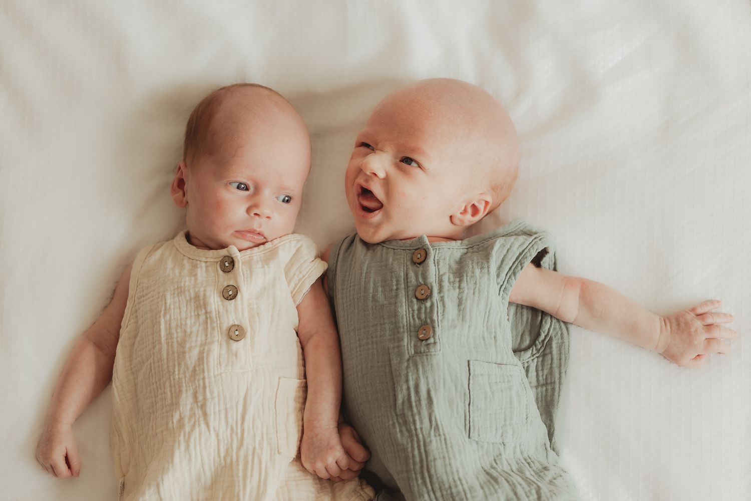 Two babies in neutral colored rompers lie together on a soft white blanket.