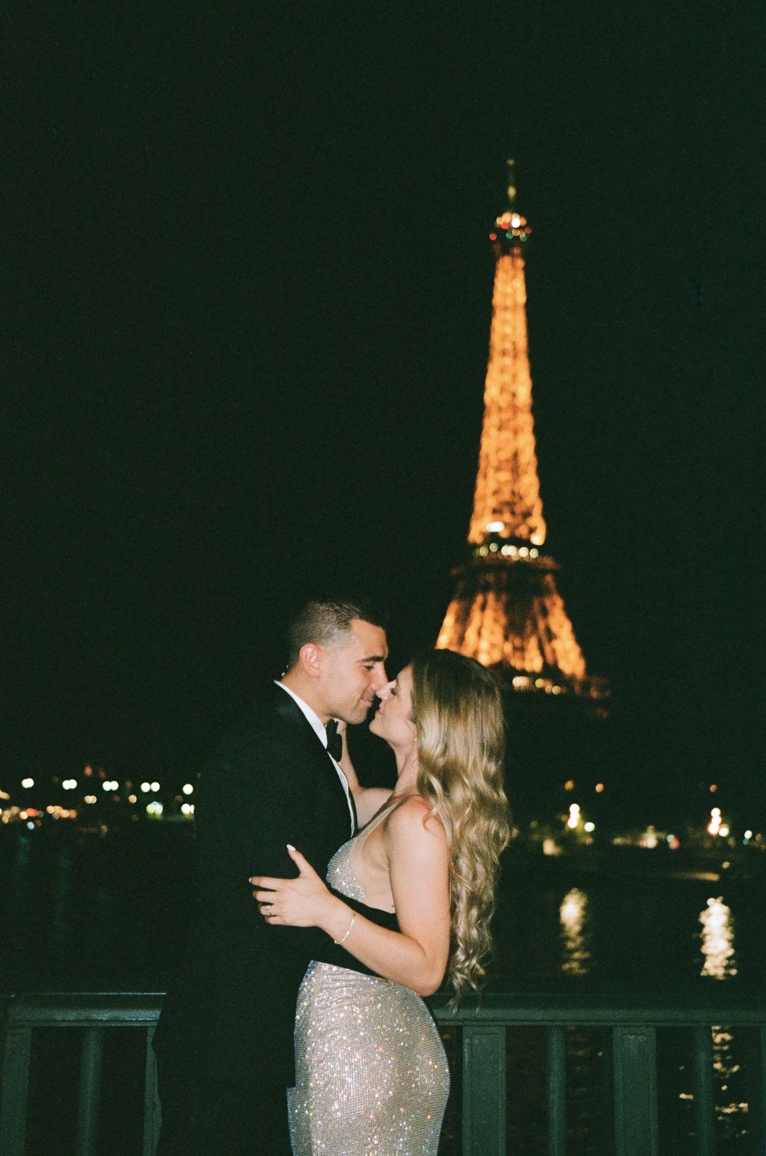 A romantic couple embraces at night with the illuminated Eiffel Tower glowing in the background across the Seine river.